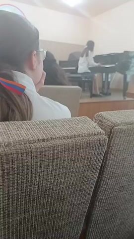 The video shows a classroom or performance space with several people seated and one person playing the piano. In the foreground, two chairs upholstered in a textured brown fabric are visible from the back. A person with dark hair, wearing glasses and a white shirt, is seated in the left chair, facing away from the camera. A colorful ribbon, appearing to be the Armenian flag colors of red, blue, and orange, is tied in their hair. Further back, another person with dark hair is seated in a chair. On a raised platform in the background, a young person wearing a white shirt is playing a black grand piano. The environment appears to be an indoor space with light-colored walls and overhead lighting. The focus is on the piano player and the audience members watching.