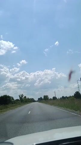 This video captures a scenic drive on a highway, likely in Alferovo, Russia, under a bright blue sky dotted with fluffy white clouds. The time of day appears to be daytime, possibly late morning or early afternoon, given the clear skies and sunlight. The road is asphalt, marked with white lines, and stretches into the distance, bordered by green grass and trees on either side. A few utility poles are visible along the roadside, suggesting a rural or semi-rural setting. There's a slight blur or smudge on the windshield, possibly a water droplet or dust, which is noticeable in the upper right portion of the frame. The overall atmosphere is peaceful and serene, evoking a sense of travel and open space. No people are visible, and the focus is on the journey itself and the surrounding landscape. There is no visible text in the video.