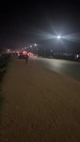A nighttime scene unfolds along a road illuminated by streetlights. A man in a patterned shirt and jeans walks along a dirt path parallel to the road. Cars are visible on the road, some with their brake lights on, suggesting a slow-moving or stopped condition. Other people are seen near the cars. The environment appears to be somewhat urban.