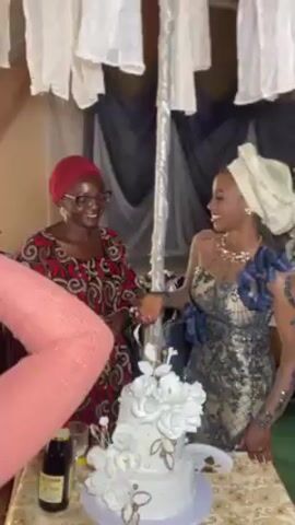 Two women stand at a table with a two-tier white cake decorated with white flowers and gold accents. The woman on the left wears a red and brown patterned dress and a red headscarf, along with glasses. The woman on the right wears a fitted dress with lace detail and a white headscarf. She holds a piece of cake and feeds it to the woman on the left. They smile at each other. Decorations hang in the background. A pink sleeve is visible in the foreground. A bottle is on the table.