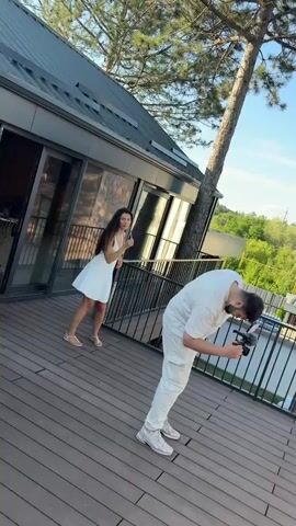 The media file appears to be a short video clip shot outdoors, possibly on a deck or balcony, with a modern house and lush greenery in the background. The time of day suggests it's late afternoon or early evening, as indicated by the warm sunlight and shadows. The weather seems pleasant, with a clear blue sky and scattered clouds.

Several people are present. In the foreground, a man in a white outfit is bent over, holding a camera and appearing to film or photograph someone. A woman with long blonde hair, wearing a black top and a white skirt, is also in the foreground, kneeling and looking towards the camera. Behind her, another woman in a white outfit is standing. Further in the background, a man in a white shirt is leaning against a railing, looking towards the camera, and another person is visible briefly. The overall atmosphere seems relaxed and social, possibly a gathering or a photoshoot. The architecture of the house suggests a contemporary style, with large windows and a wooden facade. The presence of trees and the distant view of more greenery imply a semi-rural or suburban setting. No visible text is present in the media file.