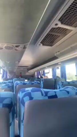 The video shows the interior of a bus with blue and grey patterned seats. The camera is positioned towards the front of the bus, looking down the aisle. A few passengers are visible, mostly from the back of their heads. The bus appears to be moving on a highway, as seen through the window. The view outside includes a road, a pedestrian overpass, and some greenery. The sky is blue and clear. The video seems to have been taken during the day.