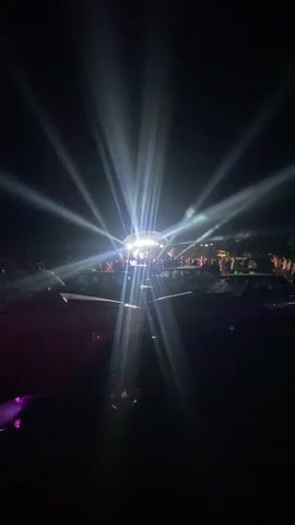 A nighttime outdoor event is depicted with bright white and purple spotlights creating beams of light that emanate from a central stage area. A crowd of people is visible in the background, gathered around the stage. In the foreground, there are dark shapes that appear to be vehicles, possibly parked cars, with their headlights or interior lights faintly illuminated. The overall atmosphere suggests a concert or a similar large-scale public gathering.
