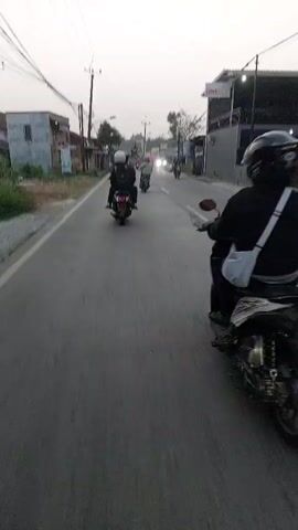 The video shows a perspective from someone riding a motorcycle on a road, possibly in Southeast Asia, given the style of the buildings and vehicles. The road is paved and appears to be a two-lane street. There are several other motorcycles on the road, some ahead and some passing. The rider in front of the camera is wearing a black jacket and a helmet. The environment is somewhat urban with buildings lining the street, some appearing to be shops or homes. Power lines are visible overhead. The sky is overcast, suggesting it may be early morning or late afternoon. The camera is positioned low, giving a clear view of the road and the motorcycles ahead. The motion of the camera indicates the rider is moving forward.