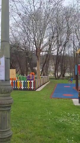 The video shows a public playground in a park. The playground is surrounded by green grass and trees with bare branches, suggesting it may be winter or early spring. Various playground equipment is visible, including a climbing structure, swings, and a small playhouse. The ground under the equipment is covered with blue and orange rubberized surfacing. A person pushes a stroller across the playground toward some swings. The sky is overcast. In the background, some buildings are visible. A lamppost stands in the foreground on the left side of the video.