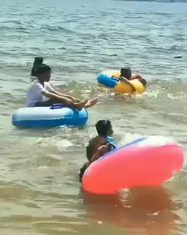 The video captures a scene at the water's edge. In the foreground, a large, inflatable pink ring floats, with a person partially visible behind it. To the left and slightly further back, a young person in a white shirt sits in a blue and white inflatable ring. To the right and further back, another person is visible in a yellow and blue inflatable ring. In the background, two dark figures are in the water, one appearing to be swimming. The water is relatively calm with gentle waves. The overall impression is of a recreational setting at a beach or lake on a sunny day.