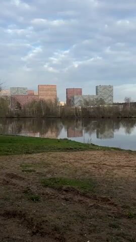 The video shows a view of a body of water with modern high-rise apartment buildings in the background. The buildings are different colors, primarily reds and grays, and are reflected in the water. The foreground is a mix of dirt, sand, and some green grass. The sky is overcast with clouds, and there are some trees along the shoreline between the water and the buildings. The camera remains still throughout the video.