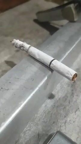 A close-up shot captures a lit cigarette resting on a metal beam. The cigarette is white with a black band near the filter. The tip of the cigarette is glowing red, and ash is accumulating. The background is out of focus, showing a concrete surface and some objects that appear to be in a workshop or industrial setting.