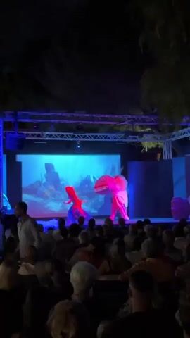 A nighttime outdoor performance is taking place on a stage in front of an audience. The stage features a large screen displaying an animated underwater scene. On the stage, a person dressed in red, presumably an actor, interacts with a large, pink, translucent creature, possibly a puppet or costume, illuminated from within. The creature appears to be a stylized representation of an aquatic animal, perhaps a manta ray or a sea serpent, with a wide mouth and flowing fins. Other performers, also in costume, are seen manipulating the creature. The audience, seated in rows, is largely obscured by darkness, with only the tops of their heads visible. Stage lighting, predominantly blue and purple, casts a dramatic glow on the performance. The overall scene suggests a theatrical or cultural event.