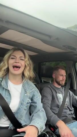 A woman and a man, wearing seatbelts, are inside a car, seemingly driving down a road. The woman is in the passenger seat, wearing a white shirt under a light-blue denim jacket. She is animatedly singing, gesturing with both hands. The man is driving, wearing a gray sweatshirt. The car is moving, evident by the changing scenery of trees visible through the car window. The car has a large sunroof. There is a phone charger connected in the car's console area.