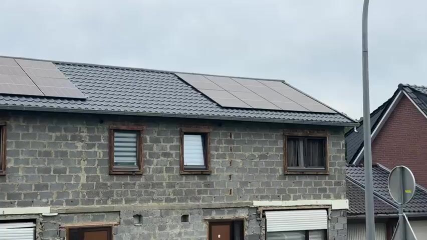 The video shows a row of attached houses, likely terraced houses, with a grey tiled roof. Some of the houses have solar panels installed on the roof. The walls of the houses appear to be constructed from concrete blocks, and some of them are unfinished or in a state of disrepair. There are windows on the upper floors, and some of the lower floors have garage doors or entrances with roller shutters. Trees and bushes are visible in front of some of the houses. The sky is overcast. The camera pans from left to right, showing different sections of the row of houses.