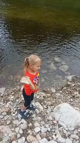 A young girl with blonde pigtails, wearing a red t-shirt and black shorts, stands on a rocky shore next to a body of water. She holds a light-colored, flat rock in her right hand. She swings her arm back, then forward, and throws the rock into the water, creating a splash. The camera follows the rock's trajectory as it enters the water and then pans slightly to show the ripples. The environment appears to be a natural setting with trees visible on the opposite bank of the water. The water is calm and appears to be relatively shallow near the shore, with rocks visible beneath the surface.