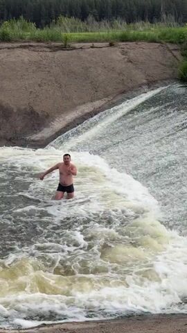 The video shows a man standing in a strong flow of water. He is shirtless and wearing dark shorts. The water is white and foamy, indicating strong currents. Behind him, a concrete structure slopes upwards, leading to a grassy area with trees in the background. The man is the focal point, positioned centrally in the frame as he faces the camera. The overall environment suggests a dam or similar water control structure.
