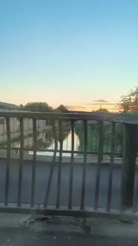 The video opens with a view of a road at dusk or dawn, with a fence and a body of water in the foreground. The sky is a clear blue with hints of orange and pink near the horizon, suggesting either sunrise or sunset. A sign that reads "BURY ROAD" is visible on the left. Trees and a distant building can be seen. As the video progresses, a black car drives down the road, followed by a white bus. A traffic light is visible on the right side of the road, mounted on a pole. The camera continues to move, showing a brick wall and more trees. The scene appears to be an urban or suburban environment.