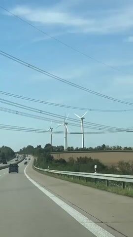 The video shows a view from a vehicle driving on a highway. Several wind turbines are visible in the distance, set against a clear blue sky with some clouds. Power lines stretch across the top of the frame. The highway has multiple lanes, and other vehicles can be seen on the road. The landscape beside the highway includes trees and fields. The camera appears to be mounted inside the vehicle, looking forward and slightly to the right.