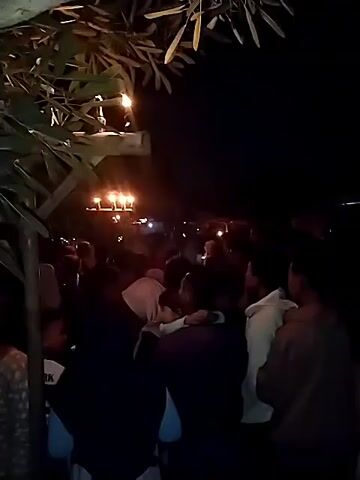 The video shows a nighttime gathering of people in an outdoor area. There are several lights visible, some overhead and some in the distance, illuminating the scene dimly. In the foreground, trees and foliage are prominent, with some branches obscuring parts of the view. A tree trunk in the mid-ground on the right has dark tape wrapped around it, securing some dark cords or wires. A crowd of people is visible in the background, appearing to be milling about or standing together. Some individuals are in focus, while others are blurred. The overall impression is of a social event taking place after dark.
