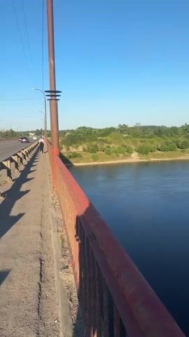 The video begins on a bridge, looking out over a wide, dark blue river. A red metal railing lines the walkway. On the left, a road with a few cars and a lone pedestrian is visible. The pedestrian appears to be walking away from the camera. The sky is a clear, bright blue. The right side of the video is dominated by the river, with a green, tree-covered bank on the far side. In the distance, across the river, a series of tall, industrial smokestacks with red and white bands are visible, emitting smoke. A smaller bridge can be seen in the far distance, connecting the two sides of the river. The camera pans slowly to the right, continuing to show the river and the distant industrial landscape.