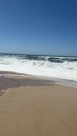 A tranquil beach scene unfolds under a clear blue sky. Gentle waves with foamy white crests roll onto a sandy shore, leaving behind a glistening sheen of water and scattered bubbles. The camera is positioned at a low angle, capturing the expanse of the ocean and the vast, cloudless sky above. The sand is a warm, tan color, with a wet patch in the foreground reflecting the sky. The waves are dynamic, with white foam swirling as they break and recede, creating a sense of peaceful motion. There are no people or man-made objects visible in the frame, emphasizing the natural beauty of the coastal environment.