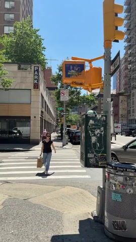 The video shows a street scene in a city, possibly New York City, with tall buildings and trees. A woman walks across a crosswalk holding a shopping bag. She is wearing sunglasses, a black t-shirt, and blue jeans. A traffic light is visible, with a sign that says "MESSIAH." A green dumpster with graffiti is next to the traffic light. In the background, cars and vans are parked on the street, and other pedestrians are visible. The weather appears to be sunny and clear.
