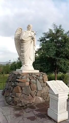 A stationary, eye-level, full-shot video captures a white stone statue of an angel standing on a circular, rough-hewn stone base. The angel is depicted with large wings, a flowing robe, and is holding a bouquet of flowers in its hands. To the right of the statue, a white stone pedestal holds an open book-like structure with inscribed text. The statue and pedestal are situated on a paved area. In the background, there are green trees and a cloudy sky. Red roses are visible on the left side of the frame. The video appears to be a slow pan or slight rotation, providing a 360-degree view of the statue and its surroundings.