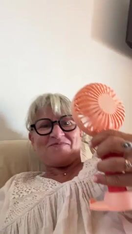 A woman with blonde hair, glasses, and a white top is sitting on a couch. She is holding a small, pink fan and moving it around. The fan is on, and it appears to be blowing air. She is smiling and looking at the camera. The background is a plain wall.