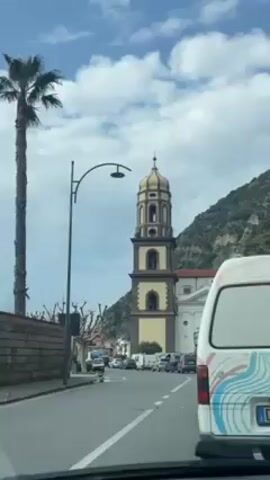 From the perspective of a moving car, a street scene is captured featuring a tall, tan and brown bell tower beside a church. A tall palm tree stands near a stone wall along the road. A decorative street light arches over the street. Various cars are parked along the street. A white van with colorful patterns on its side is visible in the foreground. A mountain rises in the background under a sky with scattered clouds.