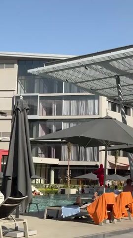 The video shows a swimming pool area at a resort on a sunny day. Lounge chairs are lined up around the pool, some occupied by people relaxing and sunbathing. Large, closed black umbrellas provide shade. There are a few palm trees around the pool.  Notable is the presence of two brightly colored sculptures: one is a green figure in a yoga pose, and the other is a yellow figure with arms raised. The architecture of the resort features a modern style, with glass windows and flat roofs. The sky is clear and blue. The music playing is dance.