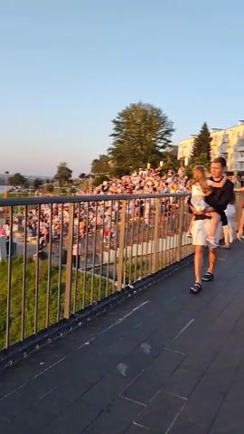 The video shows a pedestrian walkway with people walking. In the foreground, a man in a white t-shirt and sunglasses walks past, followed by a woman in a red shirt and black shorts. Further in the background, a family walks, with a man carrying a child. There are buildings in the background, and a body of water and a grassy hill are visible on the right side of the frame. The sky is clear and blue, with the sun setting. The overall atmosphere is that of a casual, outdoor gathering.