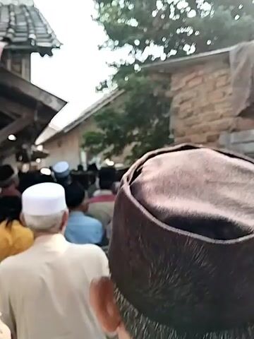 A crowd of men dressed in Muslim attire are walking down a narrow alleyway between buildings. The buildings are simple structures made of brick and wood, with tiled roofs. Some of the men are wearing white skullcaps, while others are wearing black or brown kufi caps. The alleyway is dusty and appears to be in a rural or developing area. The video is shot from behind the crowd, looking forward. The men are walking in the same direction, and it is unclear where they are going or what they are doing.