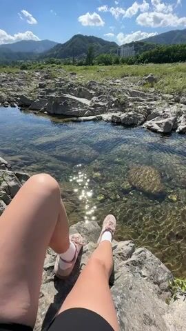The video, captured in Buk-myeon, South Korea, presents a serene, sunny daytime scene by a rocky stream. The camera is positioned from a first-person perspective, showing a person's bare legs and feet, clad in light-colored sneakers and white socks, resting on a rock.

The foreground is dominated by the clear, shallow water of the stream, which flows over a bed of grey and brown rocks. Sunlight glints brightly off the water's surface, creating sparkling reflections. Surrounding the stream are numerous grey rocks and patches of green grass and weeds.

In the mid-ground, the stream meanders through a wide, open landscape, with more rocky terrain and sparse vegetation. The background features a range of green, rolling mountains under a bright blue sky dotted with fluffy white clouds. To the far right, a faint outline of buildings suggests a distant settlement or village. The overall atmosphere is one of peaceful natural beauty on a clear, warm day.