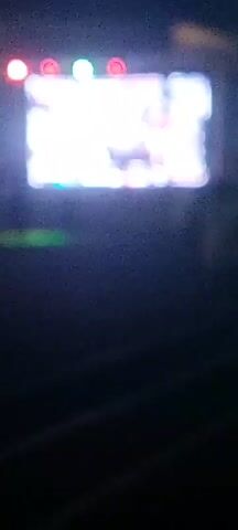 The video shows a blurry, dark scene with a large, bright screen in the background. There are several people visible in the foreground, mostly silhouetted against the light. The screen displays what appears to be a video or game with bright colors and figures. The overall atmosphere is that of a public event, possibly a concert or festival, at night.