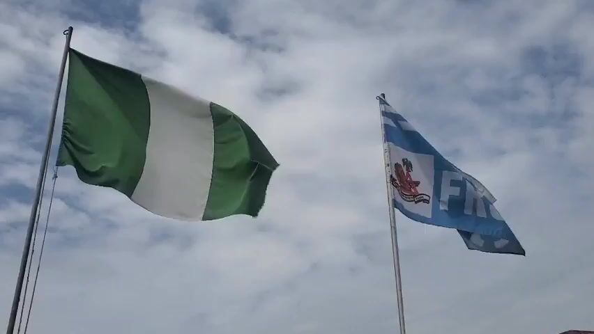The video displays two flags against a cloudy sky. The flag on the left is the Nigerian flag, featuring three vertical bands of green, white, and green. It is rippling in the wind. To the right of the Nigerian flag is a blue flag with white horizontal stripes at the top and bottom. In the center of the blue flag is a circular emblem with text and a red figure. The letters "FRSC" are prominently displayed in white on the right side of this blue flag. Both flags are attached to poles.