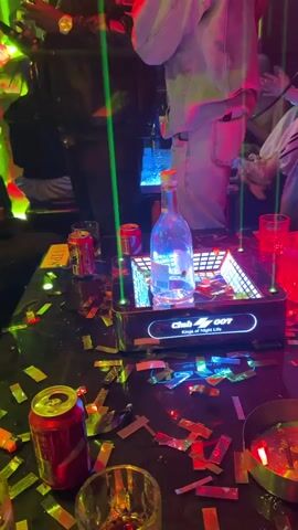 The video shows a lively scene inside a nightclub. People are standing around a table that is littered with red soda cans, empty glasses, and gold confetti. A bottle is displayed in a lit-up tray on the table. Green laser beams cut through the air. The lighting in the club changes between green and red. Individuals are holding drinks and phones and talking. One person wears a shirt with the words "RUN THE WEST" printed on the back.