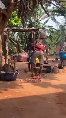 The video shows a daytime scene in what appears to be a rural African village. Several people, including children, are visible around a cooking area. In the foreground, a child in a dress and a child in a striped shirt and shorts are standing near a cooking pot on a small stool. Another person is seated on a stool nearby. The cooking area includes a traditional clay stove with pots on it. Chickens are roaming around the area. In the background, there are trees, banana plants, and a small building constructed from bamboo. The ground is covered in reddish-brown dirt. The general atmosphere suggests a simple, communal lifestyle.