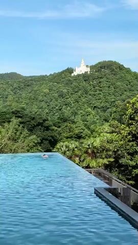 This video captures a serene scene at a resort in Ban Pong Nuea, Thailand. The focal point in the foreground is a shimmering, crystal-clear infinity pool, its surface rippling gently under a bright, sunny sky. The pool extends towards the viewer, creating a sense of depth.

In the middle ground, a single person is visible swimming in the pool, their body partially submerged, suggesting a leisurely activity. Beyond the pool, a lush, verdant landscape unfolds. Dense, emerald-green trees blanket rolling hills, creating a vibrant backdrop.

Dominating the distant background, perched atop one of the hills, is a striking white temple or structure, its architectural details standing out against the natural greenery and the clear blue sky. This prominent landmark adds a touch of cultural significance to the natural beauty of the scene. The presence of palm trees near the pool's edge hints at a tropical or subtropical climate.

The time of day appears to be midday, judging by the bright sunlight casting no distinct shadows and the clear visibility of the landscape. The weather seems pleasant and fair, ideal for outdoor activities. The overall atmosphere conveyed is one of tranquility, relaxation, and natural beauty, with a blend of man-made luxury and the splendor of nature. There is no visible text in the video.