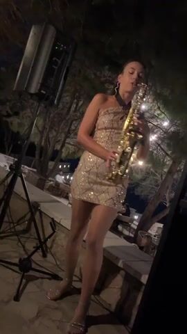 A woman plays a saxophone at night outdoors. She is wearing a sequined, one-shoulder dress and pantyhose. The saxophone is brass. She is standing on a stone ledge. A speaker is to her left, and a microphone stand is in front of her. In the background, there are trees with lights strung through them, and tables and chairs for diners. Some people are visible at the tables.