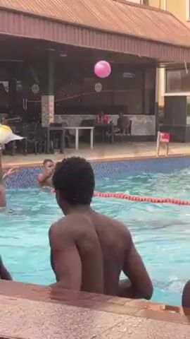 The video shows a group of shirtless young men in a swimming pool. Some are standing, some are swimming, and a few are near the edge. The pool is surrounded by buildings, including one with air conditioning units visible. There is also a covered area with tables and chairs nearby. The overall setting appears to be a hotel or resort.