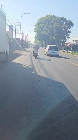 A motorcycle rider is seen from behind from a low angle, moving along an asphalt road in a forward direction. The rider is wearing a dark helmet and a white jacket. In the background, a white car is visible in the same direction. To the left of the road, there are several power lines and some buildings. Red and white flags are hanging along the side of the road. The sun is shining brightly, casting long shadows on the road. The sky is clear and blue.