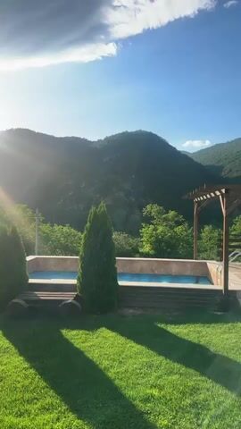 The video presents a scenic outdoor view with a swimming pool and mountains under a clear blue sky. The camera pans from left to right, revealing a small rectangular swimming pool with light blue tiles, a small evergreen tree beside it, and a wooden pergola with benches on a stone patio. Further to the right, a large trampoline with a black safety net is visible on the green grass, followed by a small, well-manicured bush. In the background, lush green mountains stretch across the horizon under a clear sky with scattered white clouds. The sun is shining brightly, casting shadows on the grass. The overall atmosphere is peaceful and serene.