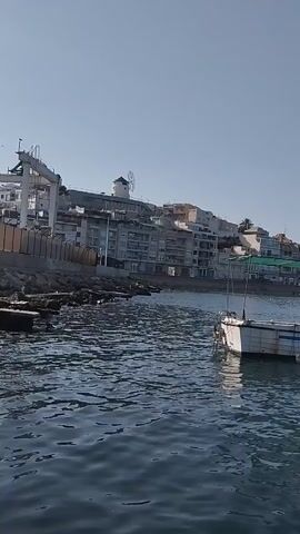 The video shows a body of water with several boats. In the foreground, a boat with a green canopy is visible. In the background, a coastline with buildings and trees can be seen under a clear blue sky. The water is slightly rippled, suggesting a gentle breeze. There are no people visible in the video.