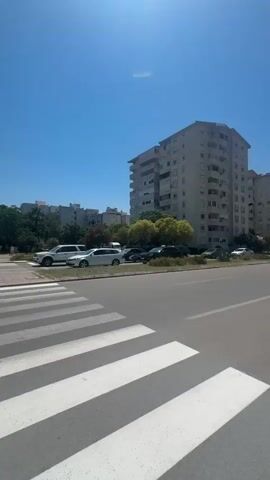 The video shows a street scene with a clear blue sky. A pedestrian crossing with white stripes is prominent in the foreground. On the right side of the crossing, a traffic light pole with red lights is visible, along with a push-button for pedestrians. Beyond the crossing, several cars are parked or driving, including a white SUV, a white sedan, and a dark-colored car. In the background, there are multi-story buildings, some with balconies, and trees. Further in the distance, mountains can be seen. A person wearing a hat and a light-colored shirt is walking on the sidewalk near the crossing. Another person is seen walking on the far side of the street. The overall impression is a typical urban environment on a sunny day.