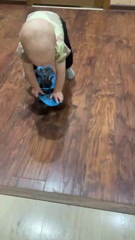 A baby wearing a camouflage shirt and black pants is on all fours on a wooden floor. The baby is holding onto a blue skateboard with black wheels, with their hands on the front of the skateboard. The baby appears to be attempting to move the skateboard. The environment is indoors, with wooden flooring and a wall visible in the background. The lighting is adequate.