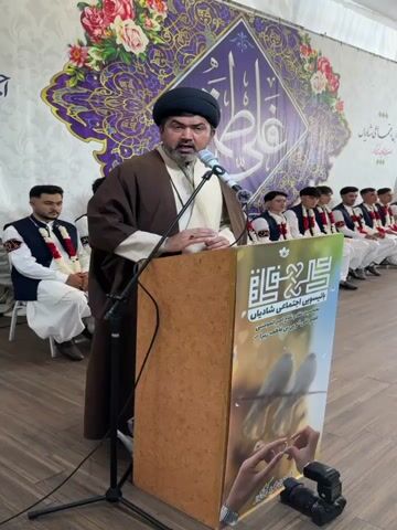 A man in a brown robe and black turban stands behind a wooden podium and speaks into a microphone. He gestures with his hand as he talks. In the background, a row of young men in matching attire sit attentively. The podium has a sign in Arabic and a picture of birds. Behind them, a large banner with floral designs is displayed. A camera is positioned on the floor in front of the podium.