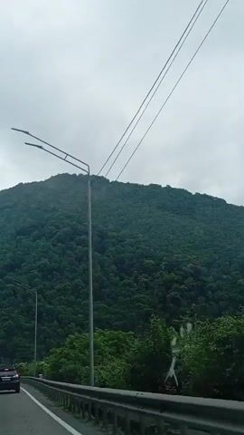 A car drives on a highway next to a densely forested mountain under a cloudy sky. There are streetlights along the highway and power lines overhead. The car in front of the camera is black.