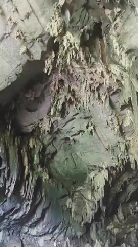 The video captures a view looking up into a large cave. Stalactites hang from the ceiling, some appearing thin and wispy, while others are thick and columnar. The cave walls are rough and textured, with visible layers of rock. In the lower part of the frame, a walkway with railings is visible, suggesting this is a tourist attraction. A few people can be seen walking on the walkway, appearing small in comparison to the vastness of the cave. The lighting is dim, casting shadows and highlighting the dramatic formations. The overall impression is of a natural wonder, showcasing the geological processes that formed the cave.