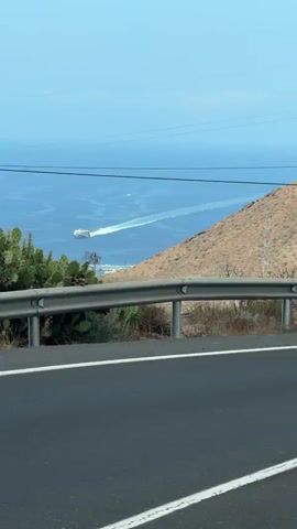 A wide shot captures a ferry leaving a foamy wake as it travels across a blue ocean. The view is from a roadside, with a metal guardrail in the foreground and a brown, rocky hillside on the right. A wooden utility pole stands near the center of the frame, with power lines stretching across the sky. In the distance, on the left side of the frame, is a low-rise building. The sky is a clear, pale blue.