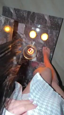 From a first-person perspective, the video shows a person sitting in a dimly lit room with a marble-topped table in front of them. The table has several lit candles, including one in a glass holder. The person is wearing a white and gray patterned robe. Their legs are visible, and their feet are near the edge of the table. The room has a calming atmosphere, suggested by the candlelight.