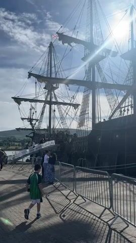 The video shows a tall ship docked at a harbor under a bright, sunny sky with some clouds. Several people are visible on the dock, including a child in a green shirt running in the foreground. Metal barriers are set up along the dock, and a ramp leads onto the ship. The ship has multiple masts and sails, and a Spanish flag is flying from the stern. In the background, another boat can be seen, as well as hills or mountains. The sun is shining brightly, creating lens flare in the footage.