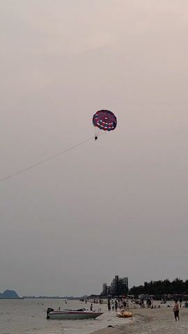 The video shows a beach scene on an overcast day. A parasailer is high in the sky, attached to a long tow line. The parachute is multicolored with red, white, and blue sections. Below, the beach is populated with people and a small boat is near the shoreline. There's also some kind of stage or construction in the background, further down the beach. The water is calm, and the overall impression is one of a leisure or vacation setting, despite the gray sky.