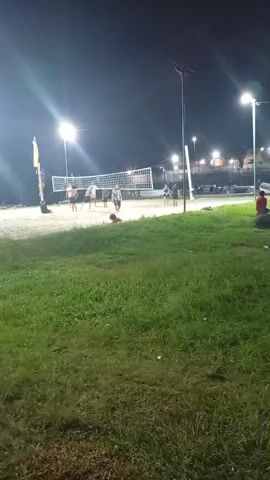 The video depicts a nighttime scene in an outdoor area, possibly a park or recreational space. In the background, a volleyball net is set up on a sandy court with several people actively playing or watching. Streetlights illuminate the scene, adding glare to the image.

In the foreground, there is a grassy area. To the right of the volleyball court, a couple of people are sitting or standing on a concrete structure. Closer to the camera, there is a worn-down slide on a small, bare patch of ground within the grass. The general atmosphere suggests a relaxed evening activity in a public space. The camera pans slightly throughout the video, offering different perspectives of the scene. The video seems to be recorded by someone standing or walking within the park.